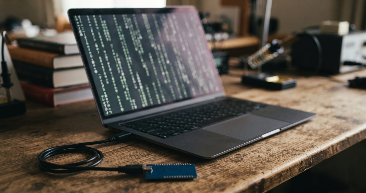 木製の机の上でノートパソコンの画面に抽象的な文字列が流れ、手前にマイコンボードとUSBケーブルが写り込んでいる写真