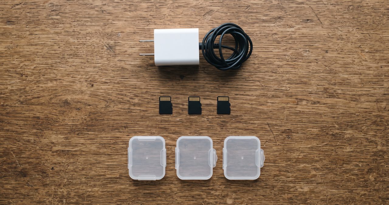 小型のACアダプター、microSDカード、透明な小型プラスチックケースを木製の机の上に種類ごとに並べた俯瞰の物撮り写真