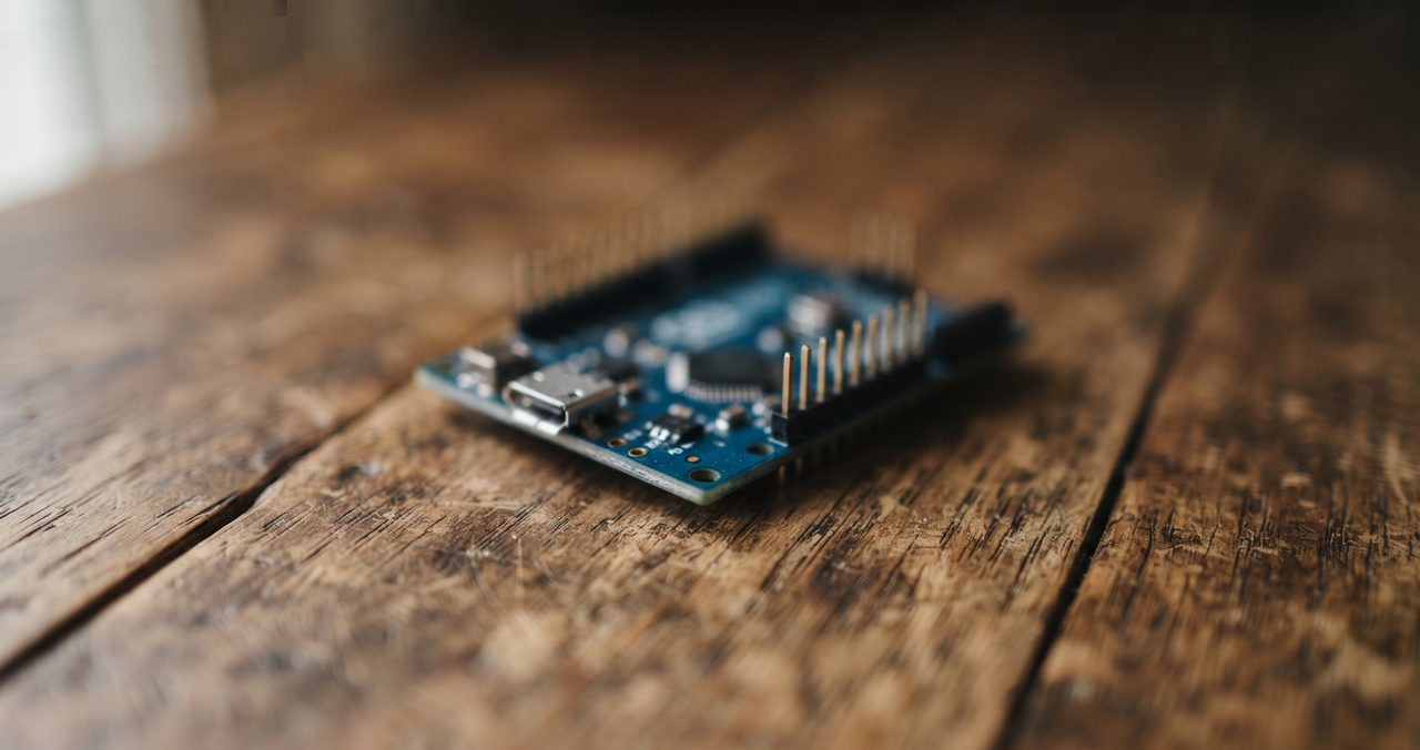 小型のマイコンボードが木製の机の上にぽつんと置かれている物撮り、基板上の文字は読み取れない角度からのクローズアップ
